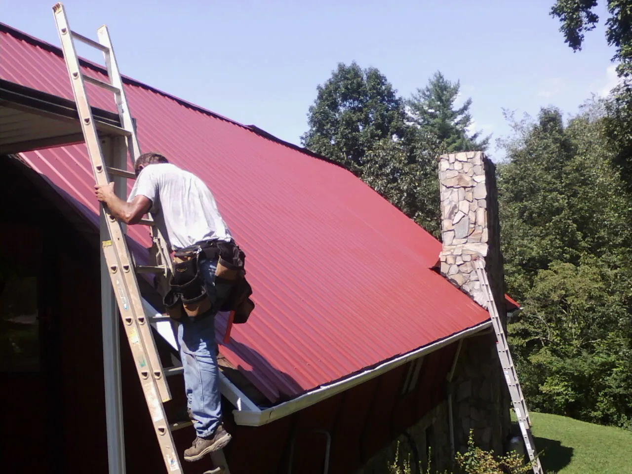 Roofer on a ladder beside a red metal roof and stone chimney — Soffit & Fascia Repair by Roofing Brothers in Vanleer